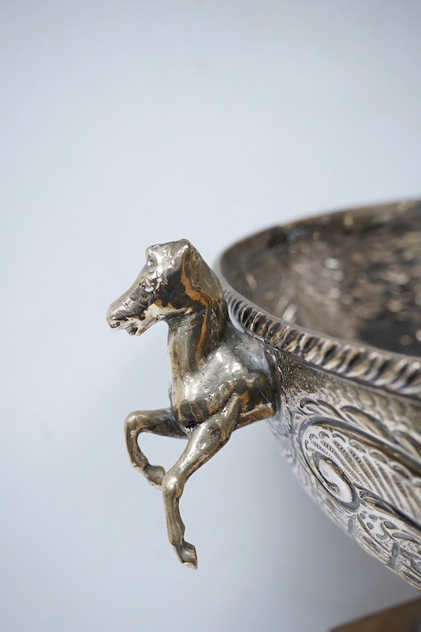 A 20th century continental white metal two handled pedestal bowl, stamped '900 SK', with horses head handles and embossed with a suspension bridge amid scrolls, diameter 28.6cm, 45.4oz, with a wooden plinth. Condition -